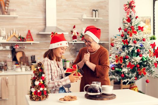 Grany looking at joygfull niece enjoying chritmas presents wearing red hat. Senior woman wearing santa hat surprising granddaughter with winter holiday present in home kitchen with xmas tree in the background.