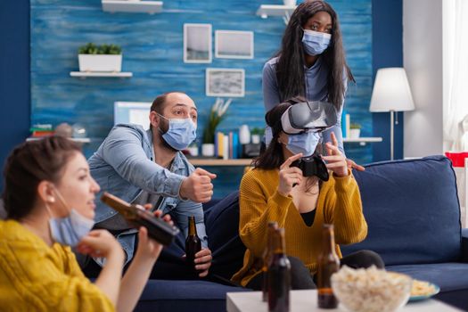 Man with face mask cheering up for friends playing video games with vr headset in home living room keeping social distancing in time of global pandemic with covid. Multi ethnic people having fun.