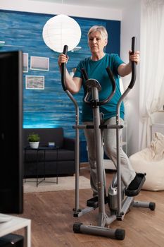 Focused senior woman working muscle legs doing body exercise using cycling bicycle machine during fitness workout. Retirement active pensioner watching aerobics video on television in living room