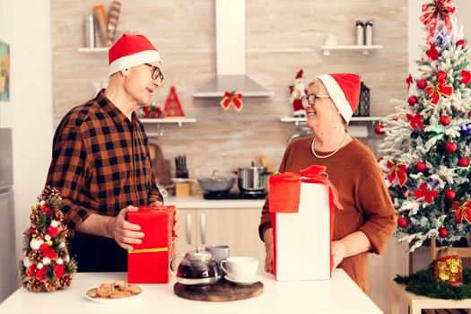 Happy grandparents celebrating christmas exchanging presents with red bow. Senior man and woman wearing santa hat during christmas giving elderly woman gift box with christmastree in the background.