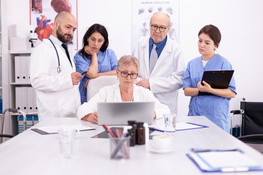 Team of doctors looking at laptop in conference room wearing medical uniform. Clinic expert therapist talking with colleagues about disease, medicine professional
