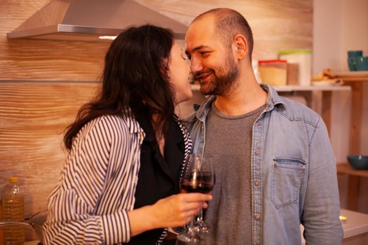 Couple touching noses and having romance date in kitchen. Adult couple at home, drinking red wine, talking, smiling, enjoying the meal in dining room.