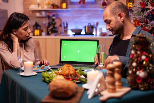 Happy family looking at green screen mock up chroma key tablet with isolated display standing on xmas decorated table. Couple celebrating christmas holiday enjoying winter season together