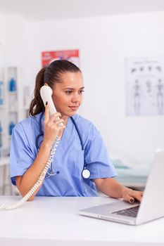 Medical practitioner having a conversation with patient on phone from hospital office and checking appointment. Health care physician sitting at desk using computer in modern clinic looking at monitor.