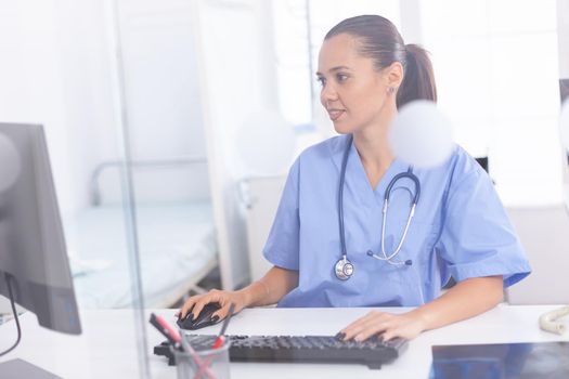 Medical practitioner using computer in hospital office. Health care physician using computer in modern clinic looking at monitor, medicine, profession, scrubs.