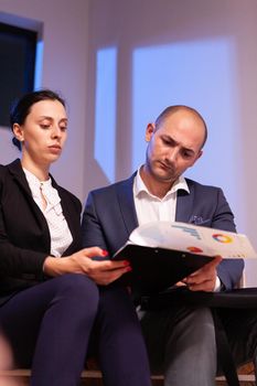 Late night after hours overworked tired businessman reading deadline raport with businesswoman doing overtime. Colleague entrepreneur working late together in evening at corporate job sitting on staircase, explaining difficult finance project.