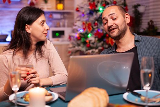 Cheerful couple shopping online xmas gift present paying with credit card on laptop computer sitting at christmas table in x-mas decorated kitchen. Happy family celebrating winter holiday