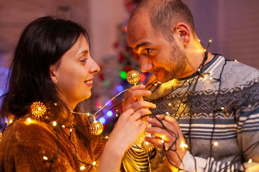 Happy amused married couple stucking in christmas tree lights enjoying spending winter holiday together. Cheerful family decorating kitchen celebrating christmastime. Santa-claus tradition
