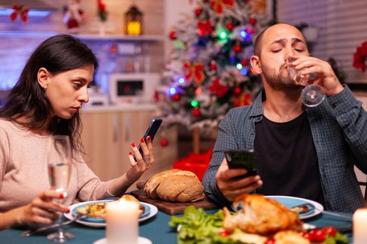 Happy family eating delicious dinner sitting at dining table in xmas decorated kitchen celebrating christmas holiday. Girfriend watching entertainment movie show using phone. Santa-claus season