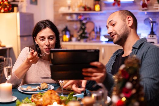 Happy family eating delicious dinner sitting at dining table in xmas decorated kitchen celebrating christmas holiday. Cheerful girlfriend reading online email using tablet computer. Santa-claus season