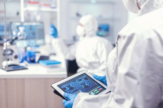 Medical engineer developing vaccine for coronavirus using tablet pc wearing ppe suit. Team of scientists conducting vaccine development using high tech technology for researching treatment against covid19
