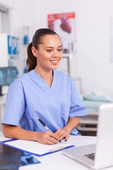 Smiling medical nurse writing notes on clipboard sitting at desk in hospital. Health care physician using computer in modern clinic looking at monitor, medicine, profession, scrubs.