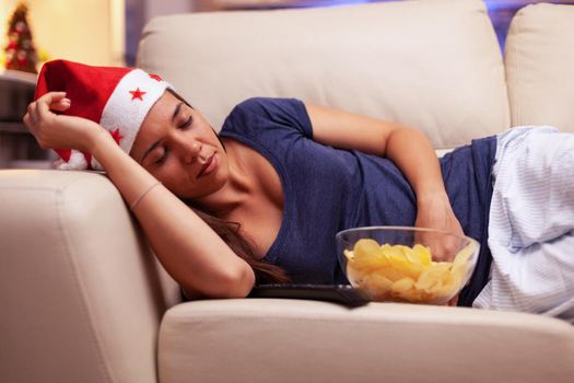 Exhausted woman falling asleep on couch while watching xmas movie on television during christmastime enjoying winter season in x-mas decorated kitchen. Adult person celebrating christmas holiday