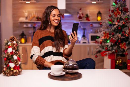 Adult woman with smartphone for video call conference on online internet and webcam with friends in kitchen decorated with ornaments and christmas lights for seasonal winter celebration