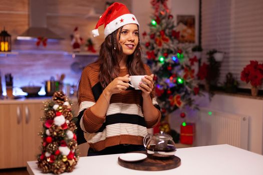 Festive woman holding cup of tea thinking about festivity in winter decorated kitchen at home. Caucasian young adult drinking tea waiting for christmas eve night celebration party
