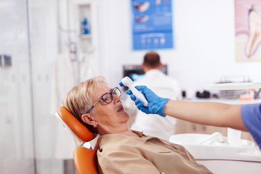 Dentist assistant holding digital body temperature indicator in front of patient forhead sitting on chair. Medical specialist in dental clinic taking patient temperature using digital device.