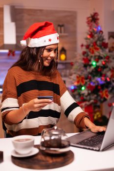 Traditional adult doing online christmas shopping for family gifts in kitchen with winter decorations, lights and ornaments for tree at home. Woman using credit card payment for purchase