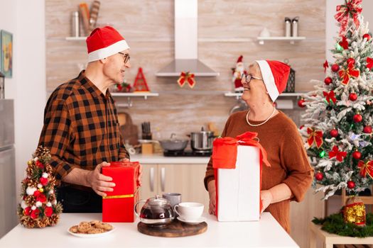 Grandparents surprising each other with xmas wrapper gift enjoying christmas holiday together in x-mas decorated culinary kitchen. Family celebrating winter season bringing present with ribbon on it