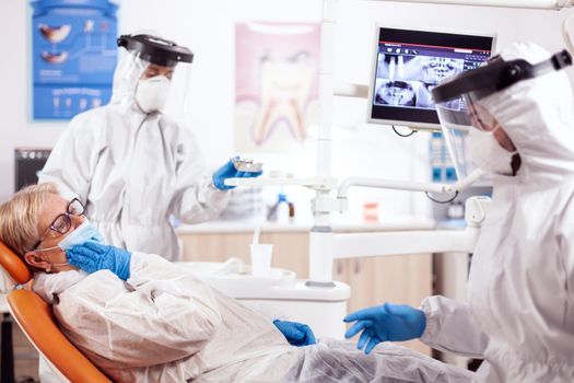 Senior patient presenting teeth pain wearing hazmat suit agains coronavirus at dentist. Elderly woman in protective uniform during medical examination in dental clinic.