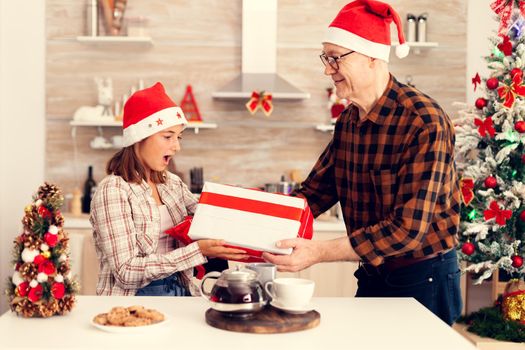 Amased cheerful niece with christmas gift box having red bow from grandfather. Senior man wearing santa hat surprising granddaughter with winter holiday present in home kitchen with xmas tree in the background.