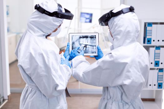 Dentistiry team in ppe suit holding tablet with teeth digital xray. Medical specialist wearing protective gear against coronavirus during global outbreak looking at radiography in dental office.