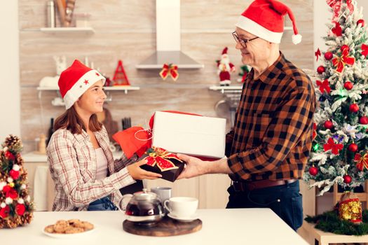 Smiling happy niece celebrating christmas with grandfather exchanging gift boxes. Senior man wearing santa hat surprising granddaughter with winter holiday present in home kitchen with xmas tree in the background.