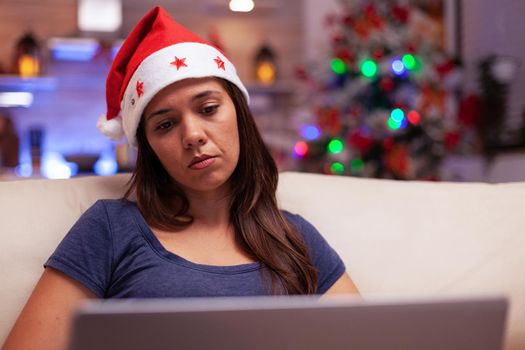 Business woman reading company email on laptop computer during christmastime sitting on sofa in xmas decorated kitchen. Girl celebrating winter holiday at home enjoying christmas season