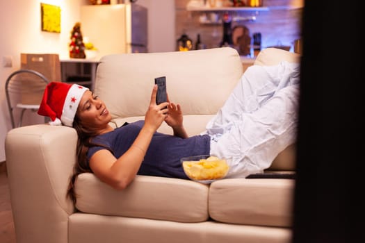 Girl messaging with friend using smartphone resting on couch in xmas decorated kitchen. Woman browsing on social media, texting during christmas holiday enjoying winter season