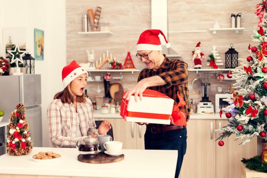 Cheerful granddad celebrating christmas giving nice gift box with red bow. Senior man wearing santa hat surprising granddaughter with winter holiday present in home kitchen with christmas tree in the background.