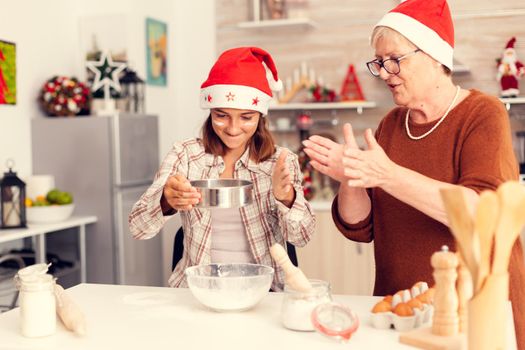 Grandmother clapping on christmas day and niece doing dough for pastery. Happy cheerful joyfull teenage girl helping senior woman preparing sweet cookies to celebrate winter holidays wearing santa hat.