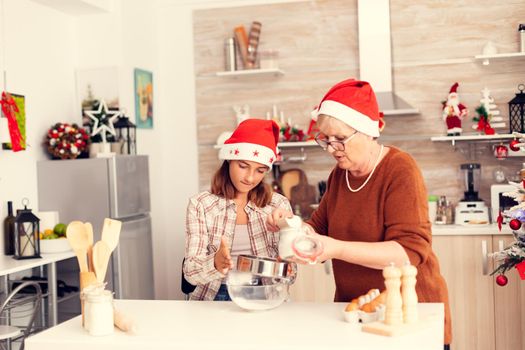 Grandmother learning niece to make delicious cookies on christmas day. Happy cheerful joyfull teenage girl helping senior woman preparing sweet biscuits to celebrate winter holidays wearing santa hat.