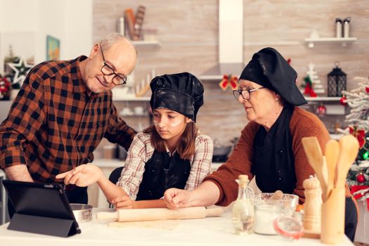 Niece and grandparents following online recipe for christmas dessert .Happy cheerful joyfull teenage girl helping senior woman preparing sweet cookies to celebrate winter holidays.