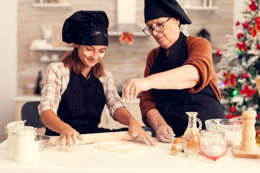 Close up of grandmother on christmas day spreading flour and making dessert with child. Happy cheerful joyfull teenage girl helping senior woman preparing sweet cookies to celebrate winter holidays.