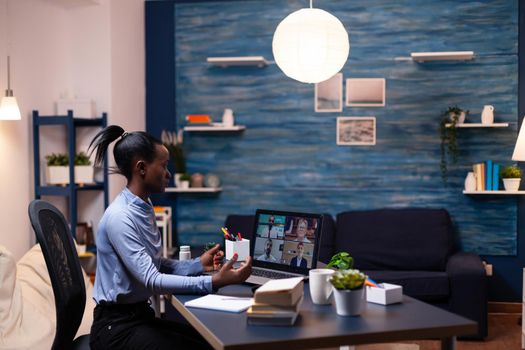 African business woman listening coworkers during video call from living room late at night. Using modern technology network wireless talking on virtual meeting doing overtime.