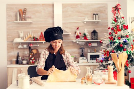 Joyful child playing with dough on christmas day wearing apron .Cheerful happy cute girl while prepearing delicious cookies for christmas celebration in kitchen with christmas tree in the background.