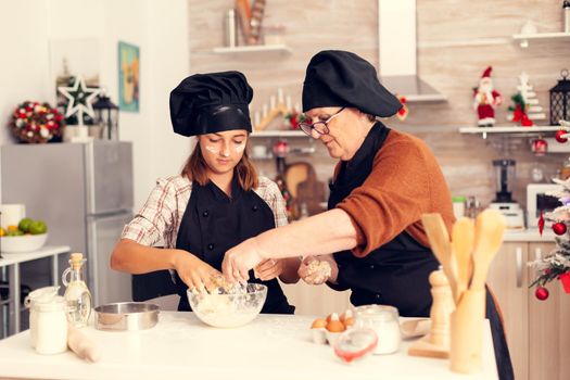 Kid making traditional cookies on christmas day wearing apron and bonette. Happy cheerful joyfull teenage girl helping senior woman preparing sweet biscuits to celebrate winter holidays.