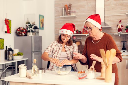 Joyfull niece on christmas day cracking eggs over flour. Happy cheerful joyfull teenage girl helping senior woman preparing sweet cookies to celebrate winter holidays wearing santa hat.