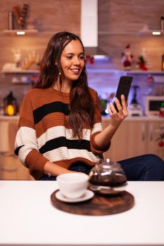 Happy woman holding smartphone for video call chatting conference on online internet with relatives in kitchen decorated with ornaments, lights and christmas tree for winter celebration