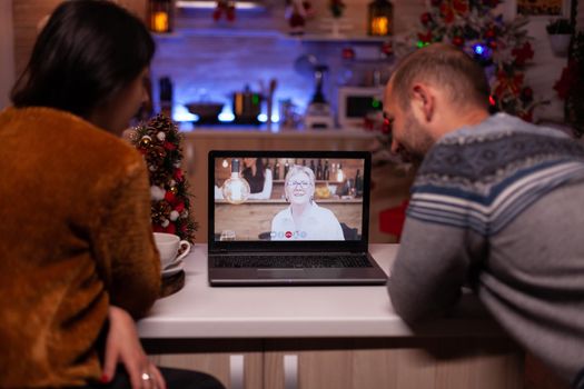 Happy family discussing with remote grandparent during online videocall conference celebrating christmas holiday together in xmas decorated kitchen. Joyful couple enjoying winter season