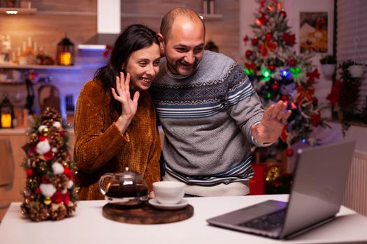 Happy family greeting friends during online videocall meeting on laptop computer celebrating christmas holiday together. Cheerful couple enjoying spending winter season in xmas decorated kitchen