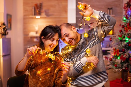 Family stuck in christmas tree lights in xmas decorated kitchen celebrating christmas holiday. Happy couple making funny while enjoying spending winter season together. New-year festive tradition