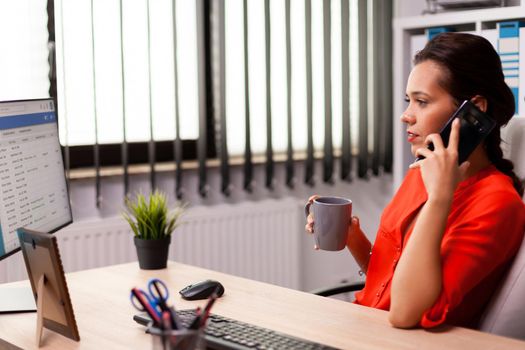 Corporate businesswoman in workplace talking on phone with business partner wearing red. Busy freelancer working using smartphone from office to talk with clients sitting at desk looking at document.