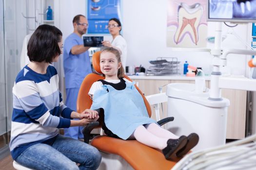 Little girl missing milk tooth wearing dental bib discussing with parent at dentist consultation. Child with her mother during teeth check up with stomatolog sitting on chair.