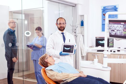 Stomatolog holding x-ray of senior woman sitting on orange chair in dentist cabinet. Medical teeth care taker holding patient radiography on tablet pc near patient standing up.