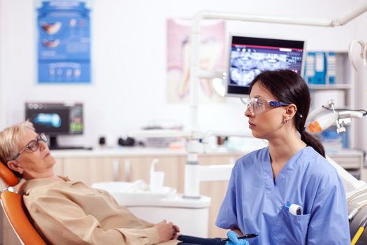 Assistant in dentist cabinet during elderly woman oral hygiene check up sitting on chair. Senior woman talking with medical nurse in stomatology office about teeth problem.