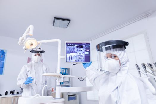 Pov of dentist and nurse in protective suits explaining teeth ex-ray on monitor. Stomatology specialist wearing protective suit against infection with coronavirus pointing at radiography.