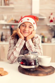Adorable teenage girl with santa hat smiling at camera celebrating christmas.Cheerful happy adorable teenager girl in home kitchen with delicious biscuits and xmas tree in the background celebrating winter holiday.