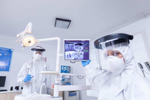 Patient pov of dentist in coverall pointing at monitor with teeth x-ray during Stomatology specialist wearing protective suit against infection with coronavirus showing radiography.