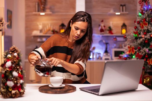 Caucasian woman pouring cup of tea from kettle while talking to friends on video call app. Young adult using laptop for christmas eve in holiday decorated kitchen with ornaments