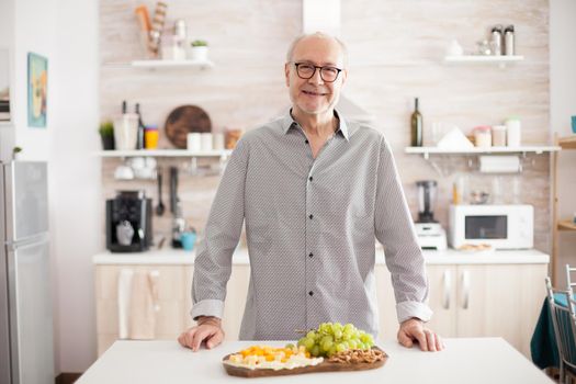Smiling senior man in home kitchen looking at camera with variety of tasty cheese and grapes on table top.